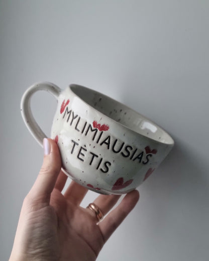 Hand holding a ceramic mug with text and heart designs against a plain background