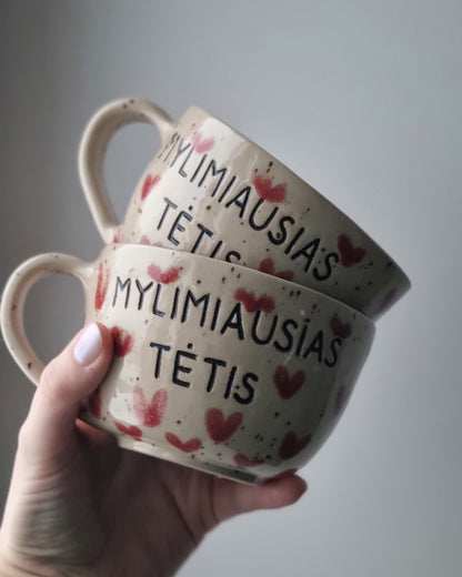 Two ceramic bowls with decorative text and heart patterns held by a hand against a plain background.